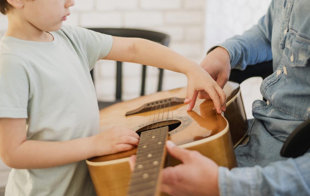 Ein Kind lernt Gitarre, daneben eine Lehrerin, die die Griffhaltung zeigt.